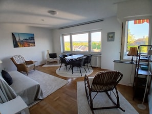 A room with a sofa, a dining table with chairs, a TV on a stand, and a window with a view of the countryside.