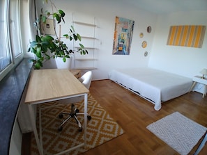 Een kamer met een houten bureau, een witte stoel, een bed, een potplant, een wandplank en een kamerbreed tapijt met een patroon.
