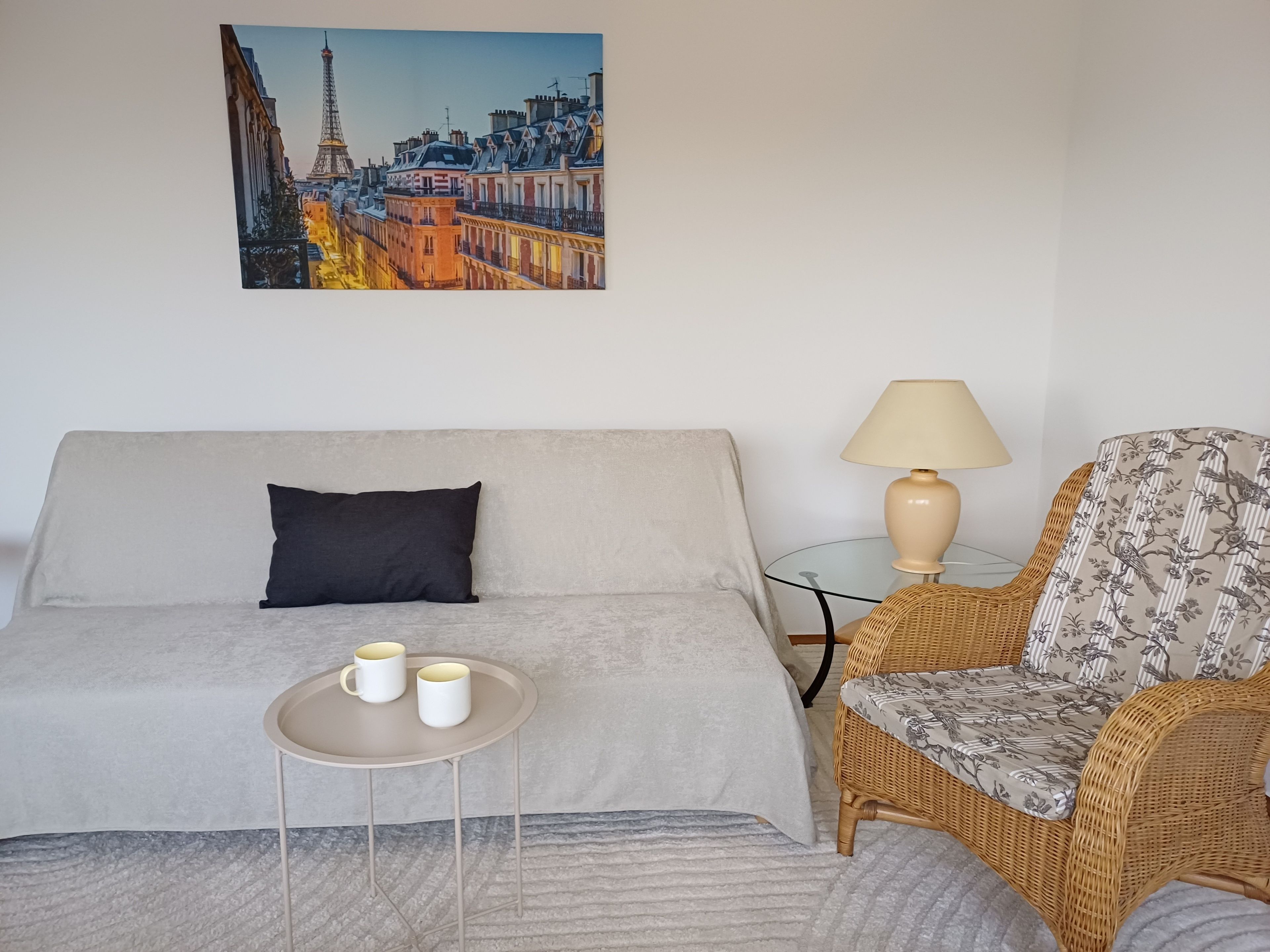 A hotel room with a bed, a small table with two cups, a wicker chair, a lamp, and a painting of a cityscape.