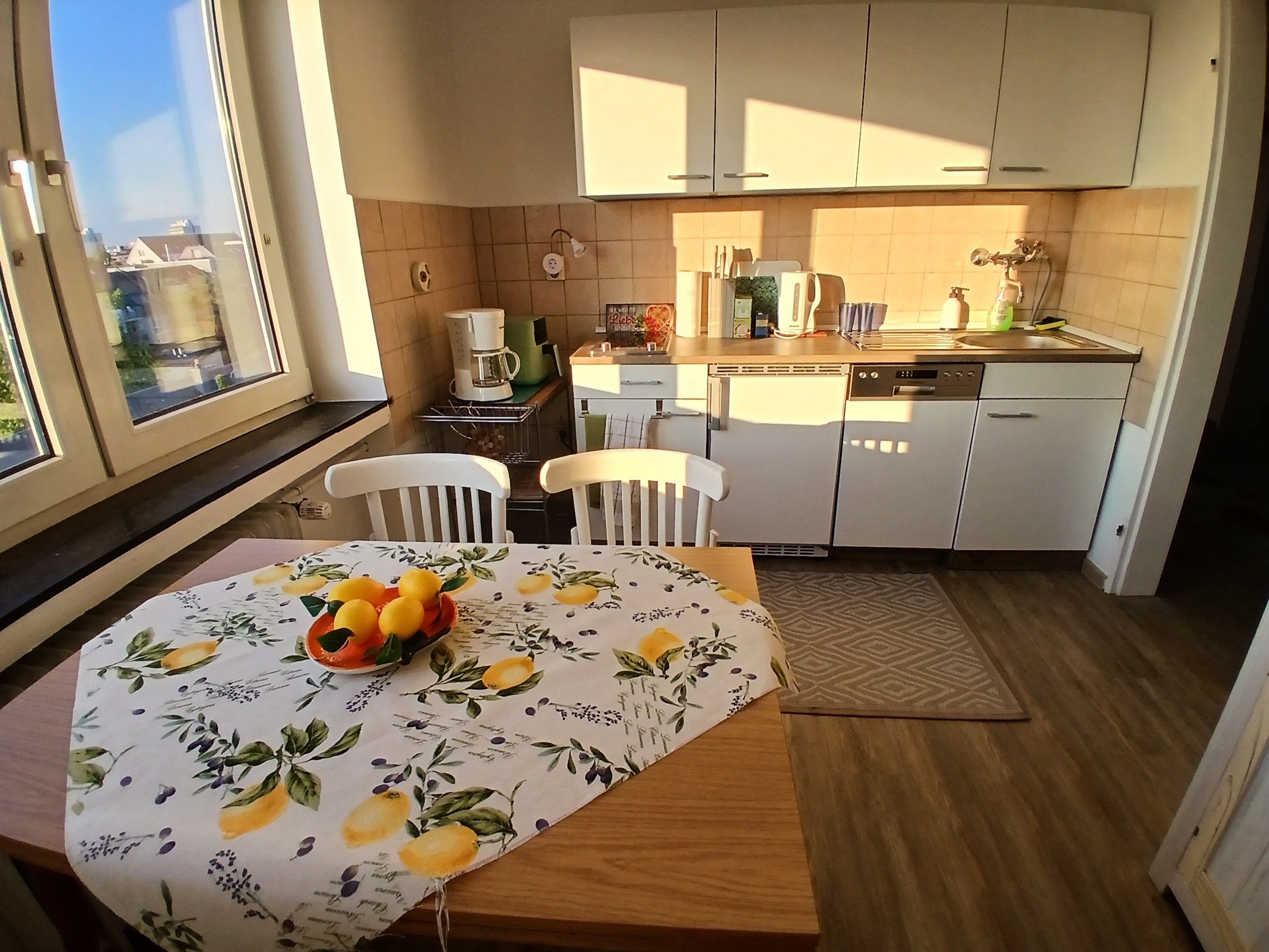 Una cocina con armarios blancos, una mesa de comedor con un centro de frutas y vistas por una ventana.