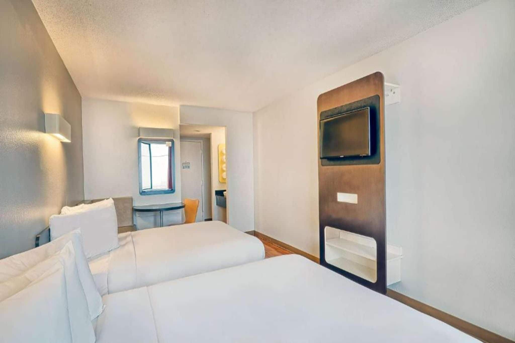 A hotel room with two beds, a TV mounted on the wall, and a small table with a chair.