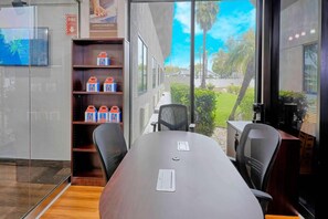 Un salón de conferencias con una larga mesa, sillas, una estantería con cajas y vistas al exterior.