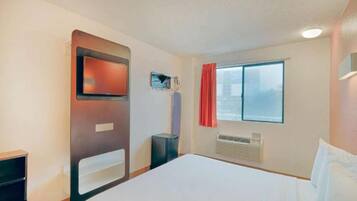A hotel room with a bed, a television mounted on the wall, a window with curtains, and an air conditioning unit.