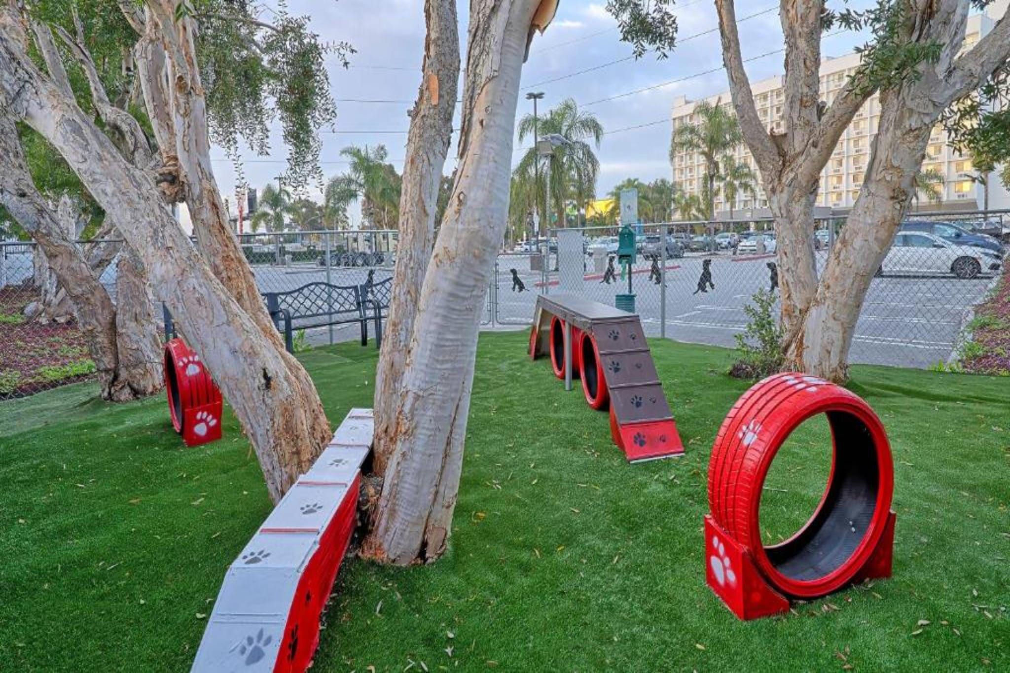 Een park met kunstbomen, een omheind gebied en een speeltuin met rode en grijze speeltoestellen.