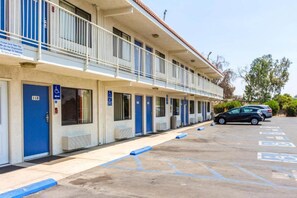Un motel avec une rangée de chambres, chacune dotée d’une porte bleue et d’une entrée accessible aux personnes à mobilité réduite. Un parking est disponible, avec des places de stationnement réservées aux personnes handicapées.