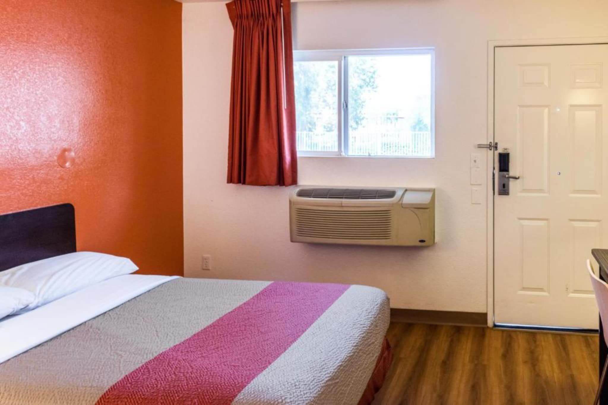 A hotel room with a bed, an air conditioning unit, a window with curtains, and a door.