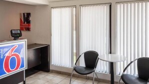 Ein Hotelzimmer mit einer Rezeption, einem digitalen Bezahlsystem, zwei Stühlen, einem kleinen Tisch und großen Fenstern mit Jalousien.