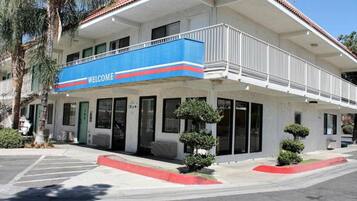 Un edificio de dos pisos con un letrero azul que dice 'WELCOME', un porche cubierto y un pequeño árbol en el jardín delantero.