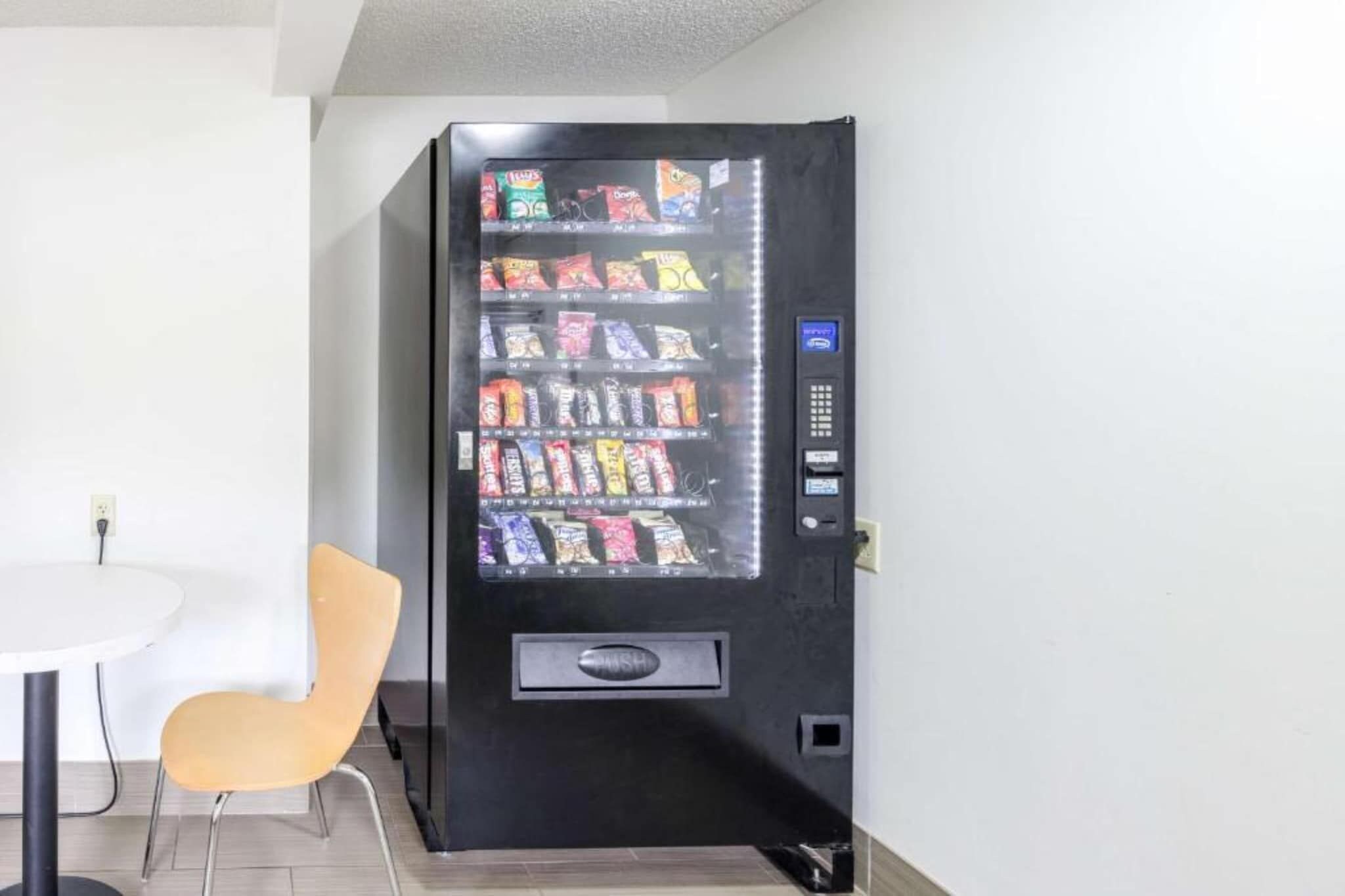 A vending machine with various snack options in a room with a white table and a single chair.