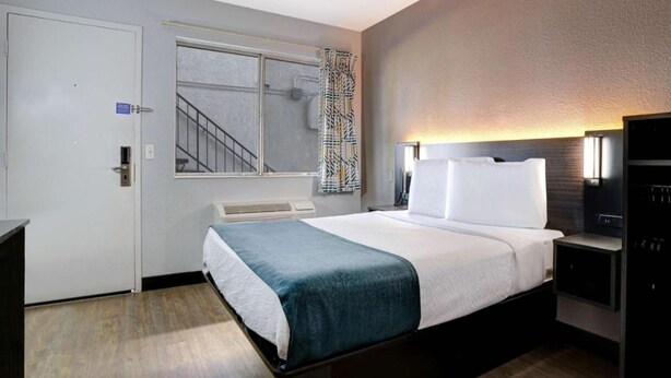 A hotel room with a bed, bedside tables, a window with a view of a staircase, and a door with a sign. - 1 Full Bed | ADA/RI Shower (Bakersfield)