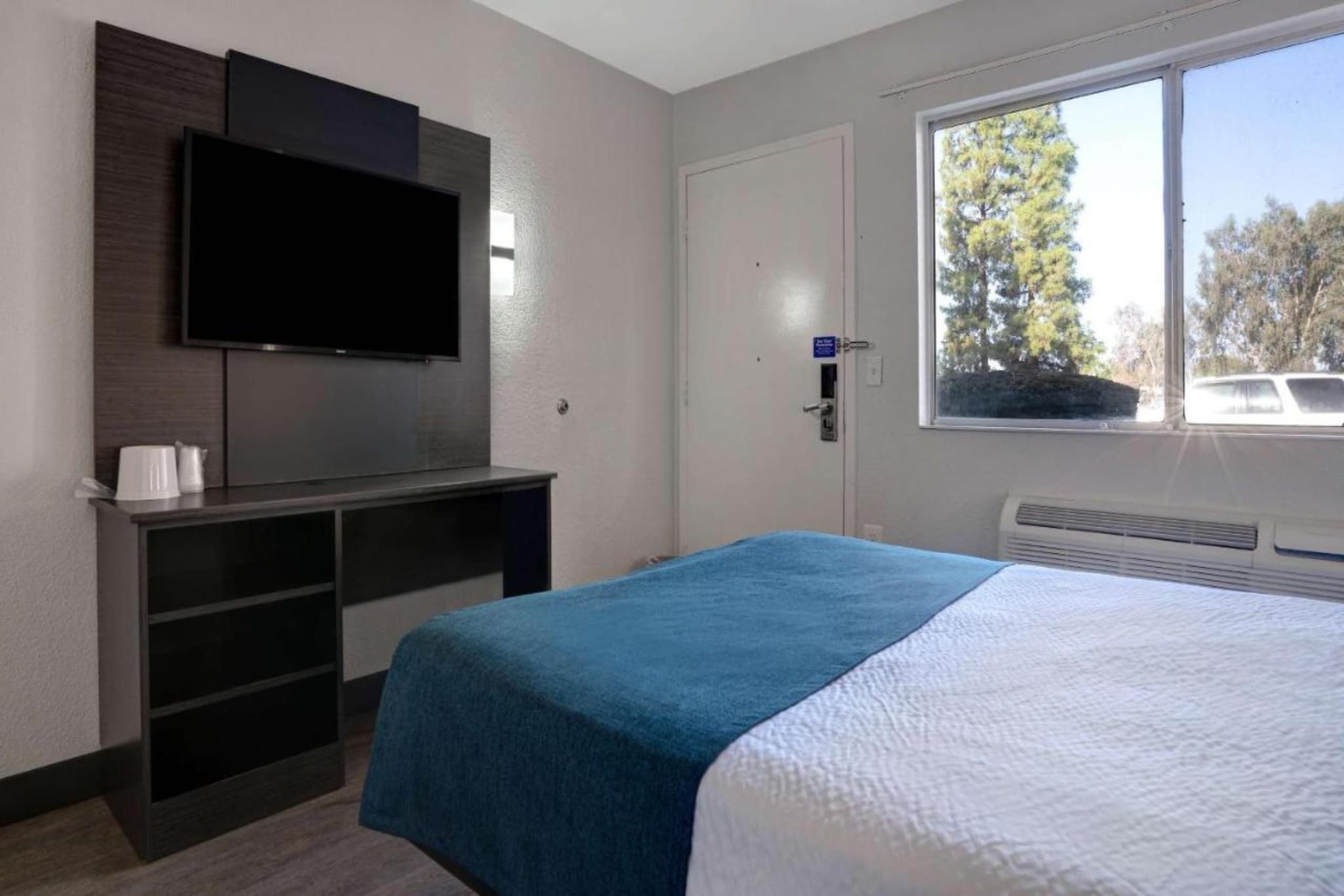 A hotel room with a bed, a TV mounted on the wall, a desk with a chair, and a window with a view of trees.