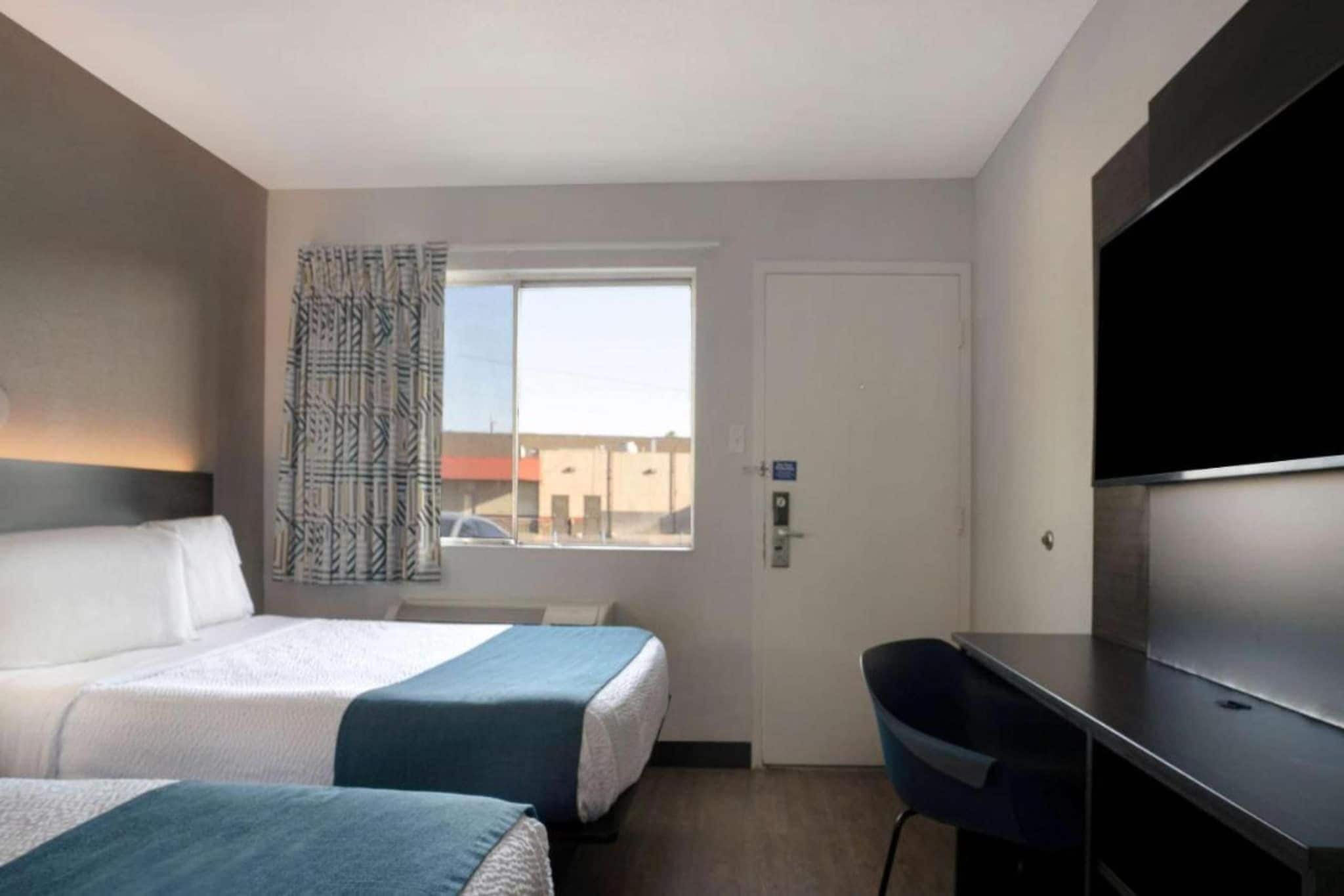 Habitación de hotel con dos camas, un escritorio, televisor y ventana con vista a edificios.
