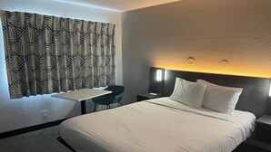 A hotel room with a large bed, a desk, a chair, and a patterned curtain. - Cozy 1 Queen Bed | Smoking (Bakersfield)
