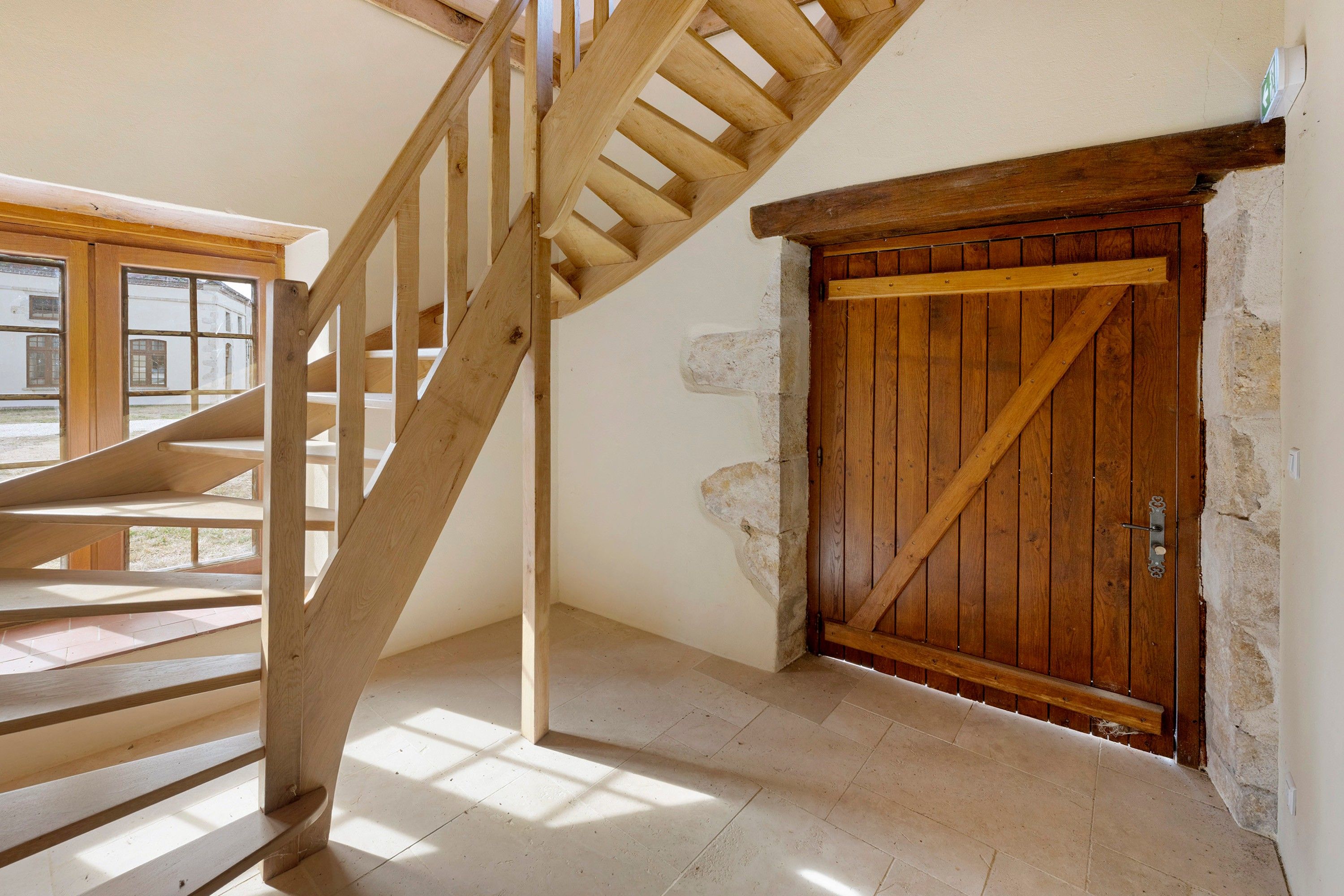 A wooden staircase leading to an upper floor, a rustic wooden door, and a view of the outside through a window.
