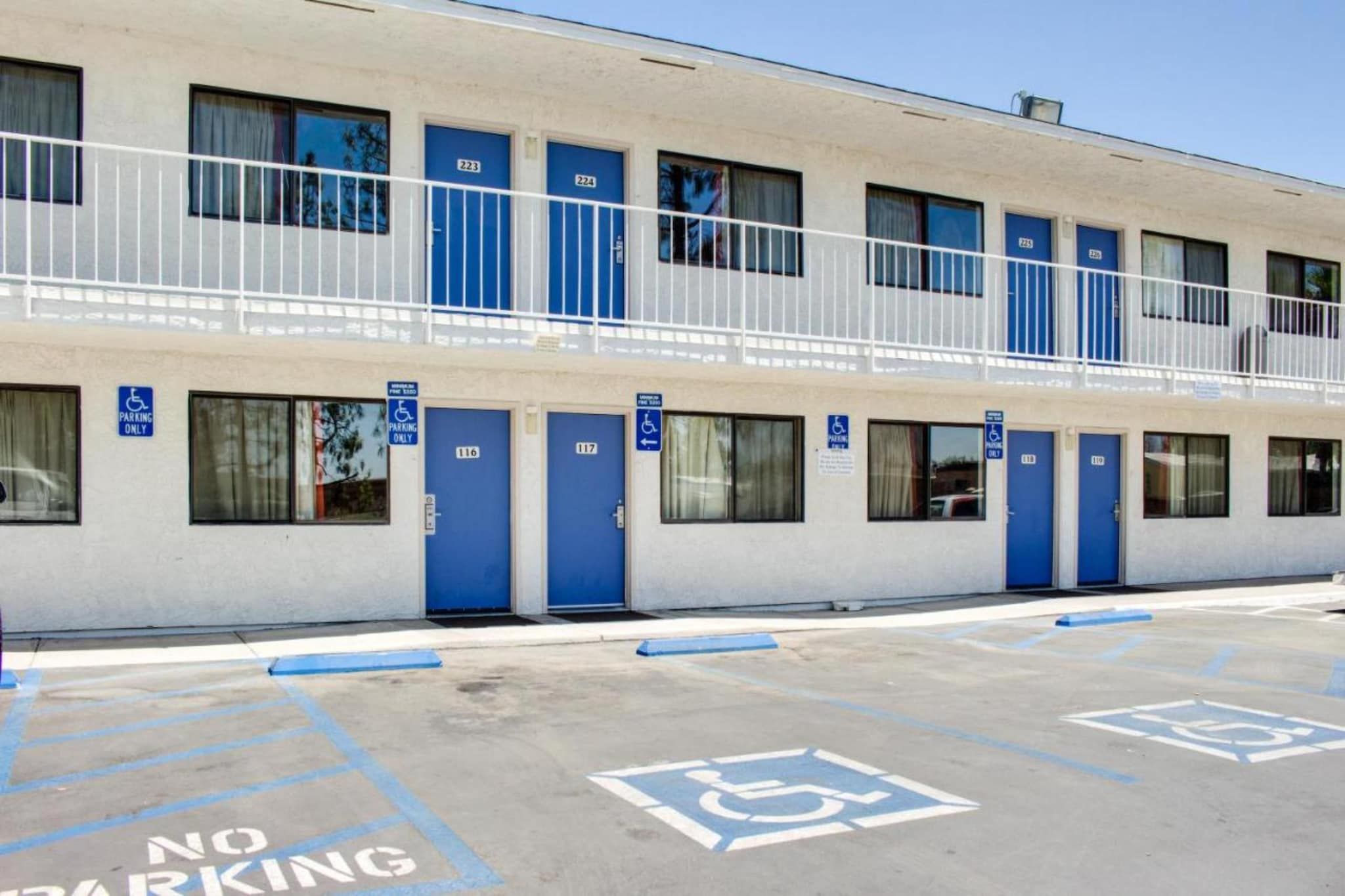 Un immeuble à plusieurs étages dont les portes sont numérotées et peintes en bleu, et dont les places de stationnement sont signalées par des panneaux indiquant qu’elles sont réservées aux personnes handicapées.