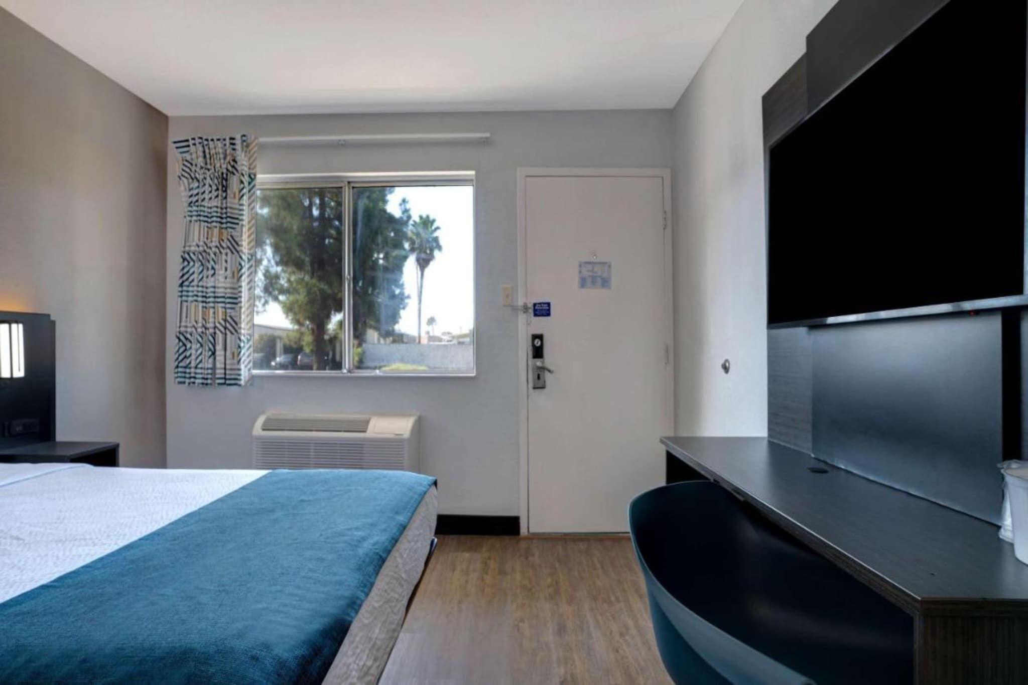 Habitación de hotel con cama, escritorio, televisor de pantalla plana, ventana con vista a palmeras y aire acondicionado.