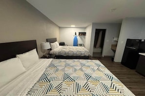 Habitación de hotel con una cama grande, una cama más pequeña, un escritorio con lámpara, una tabla de planchar azul y un refrigerador negro.