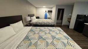 Habitación de hotel con una cama grande, una cama más pequeña, un escritorio con lámpara, una tabla de planchar azul y un refrigerador negro.