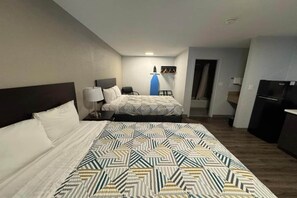 Ein Hotelzimmer mit einem großen Bett, einem kleineren Bett, einem Schreibtisch mit Lampe, einem blauen Bügelbrett und einem schwarzen Kühlschrank.