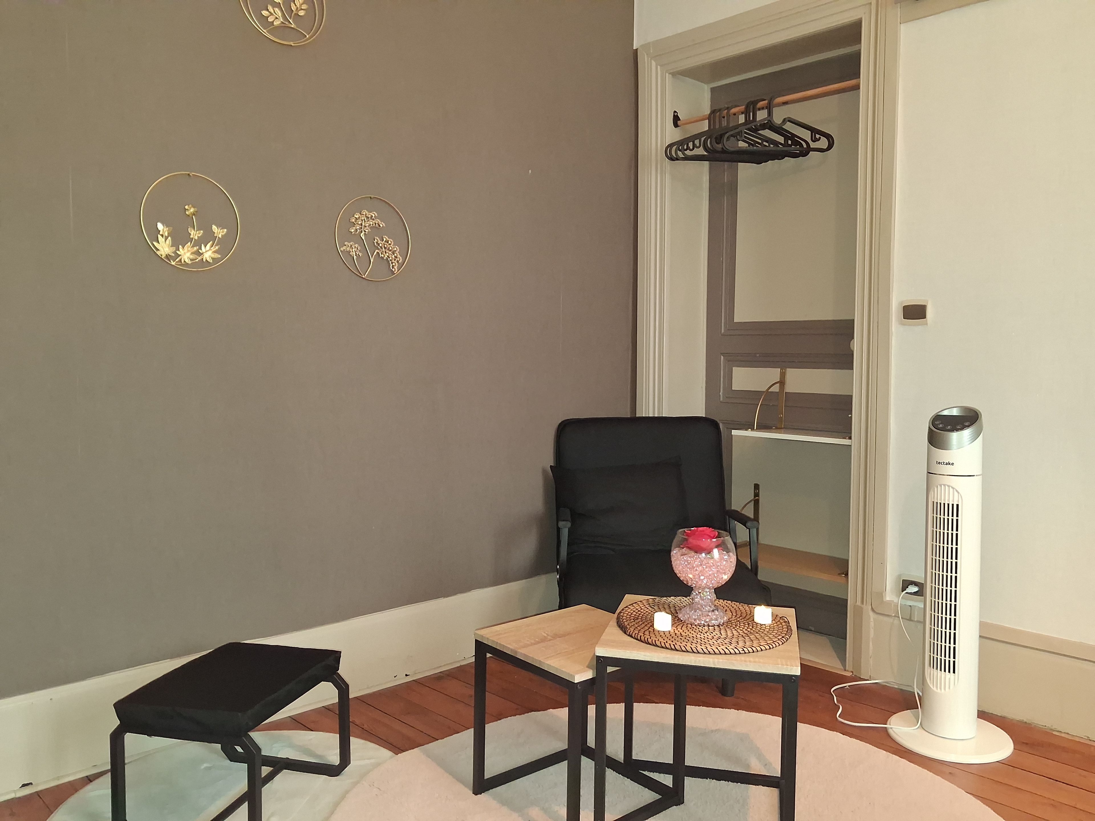 A room with a black chair, a small table with a decorative item, a standing heater, and a coat rack.