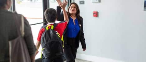 Una persona con camisa azul y chaqueta negra caminando hacia una puerta con un botón de emergencia rojo en la pared.