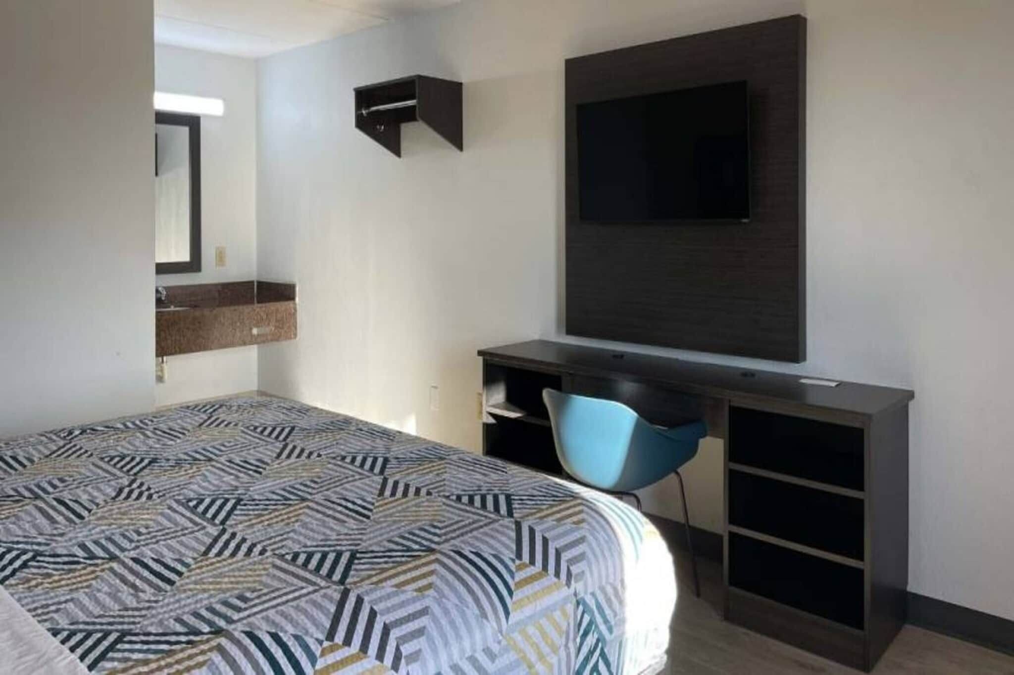 Una habitación de hotel con cama, escritorio con silla, televisor y estante en la pared.