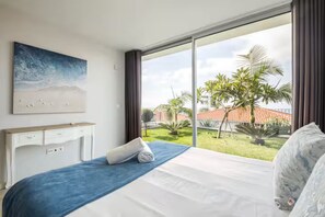 Une chambre d’hôtel avec un lit, un bureau et une grande fenêtre donnant sur un jardin et des palmiers.