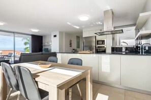 Une cuisine moderne avec des armoires blanches, un coin repas doté d’une table et de chaises en bois, et une vue sur l’océan grâce à de grandes fenêtres.