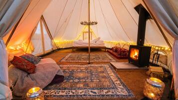 A tent interior with a central bed, a wood-burning stove, and decorative lighting.