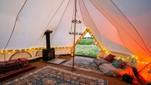A cozy tent interior with a central fire pit, rugs, and a view of the outdoors.