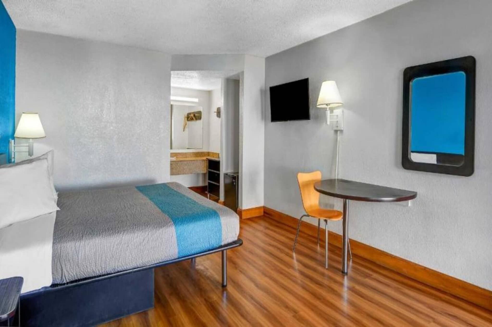 A hotel room with a bed, a desk, a chair, a flat-screen TV, and a wall-mounted lamp.
