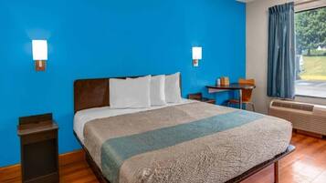 A hotel room with a large bed, a desk, and a chair. There is a window with curtains and an air conditioning unit.