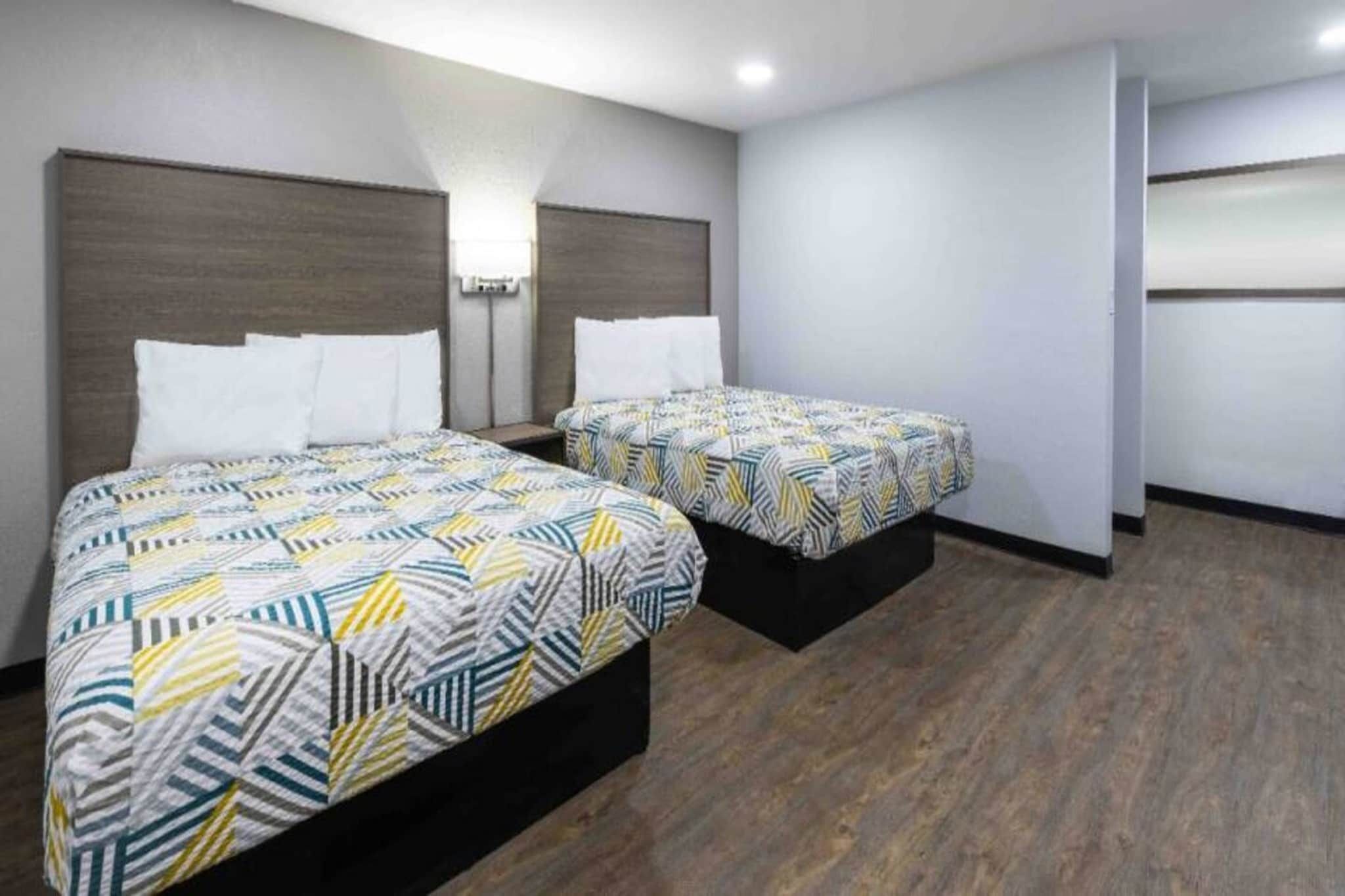 Una camera d'albergo con due letti, ciascuno con una coperta a motivi e un poggiatesta in legno, oltre a un comodino con una lampada.