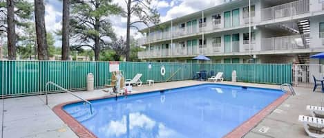 Une piscine bordée de briques, entourée d’une clôture verte, et un immeuble à plusieurs étages avec des balcons en arrière-plan.