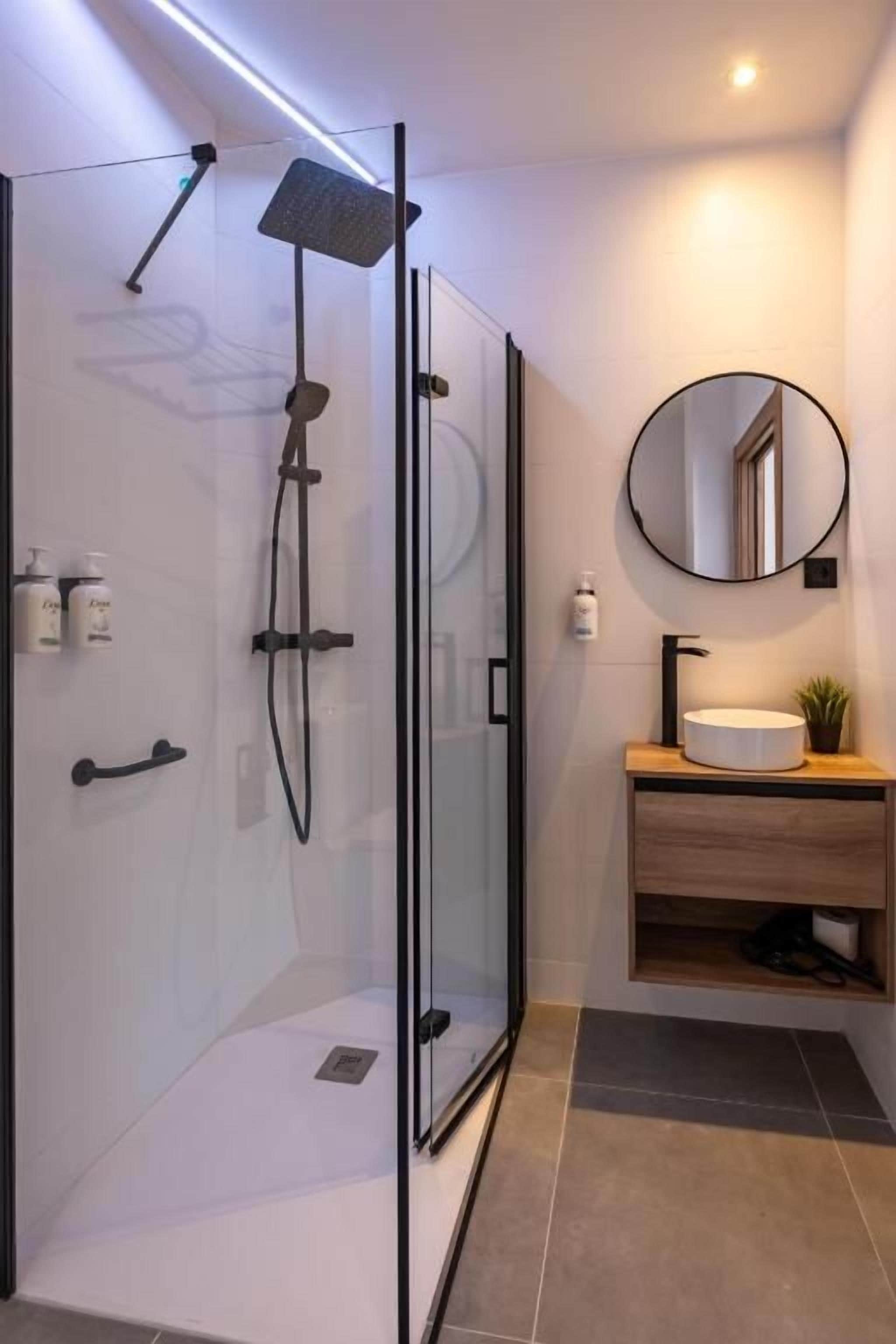 Un baño moderno con ducha de vidrio, un espejo redondo y un tocador de madera.