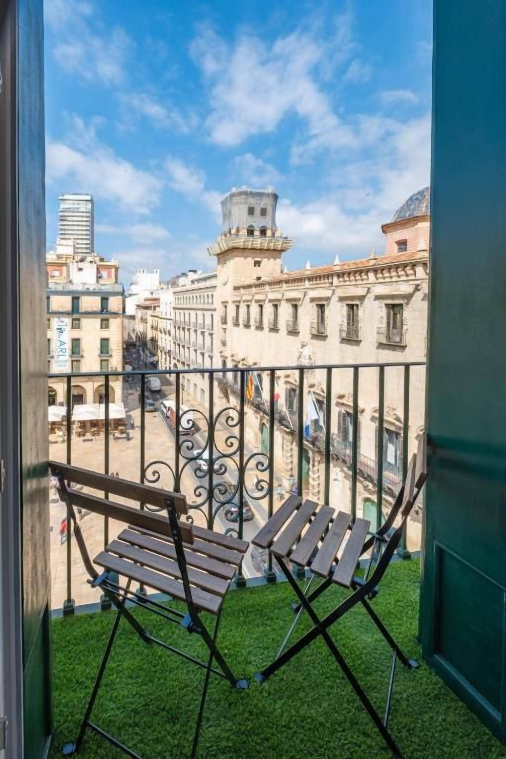 Un balcón con dos sillas de madera, barandilla de hierro forjado y vistas a edificios históricos.