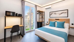 Quarto de hotel com cama, escrivaninha, cadeira, guarda-roupa e uma janela grande com vista para edifícios.