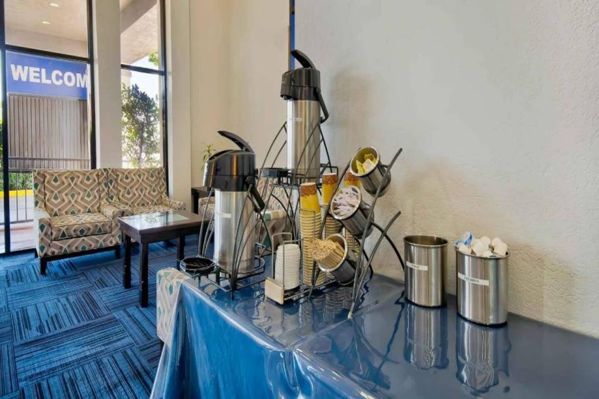 Ein Hotel-Empfangsbereich mit einer Kaffeestation, einer Couch und einem 'WELCOME'-Schild.