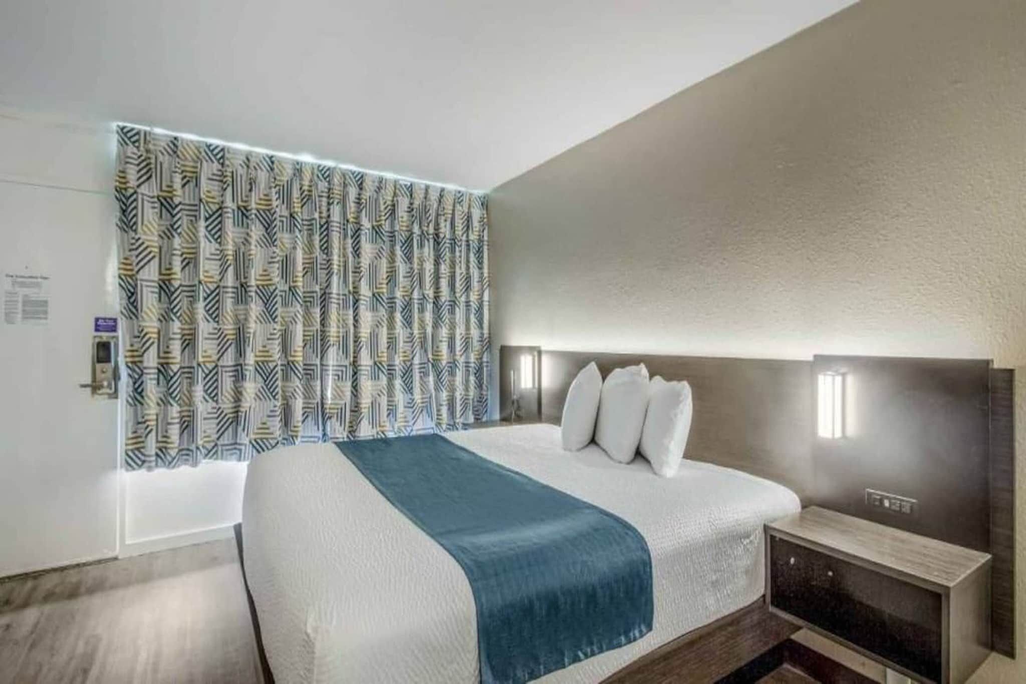 Moderni hotellihuone, jossa on suuri sänky, sängynpääty, jossa on integroitu valaistus, yöpöytä ja kuviollinen verho.