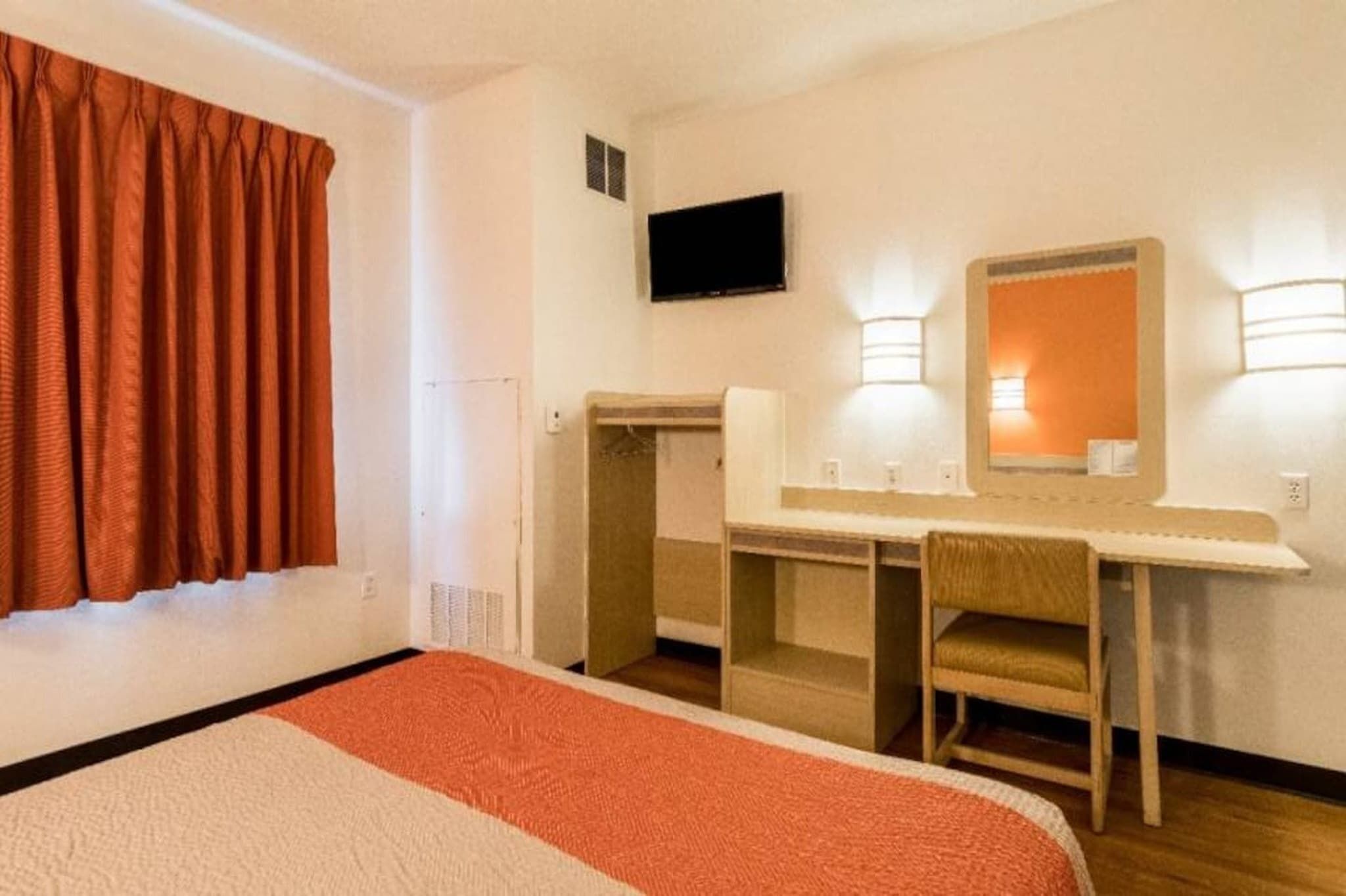 Habitación de hotel con una cama, un escritorio, una silla, un televisor y dos luces fijadas en la pared.