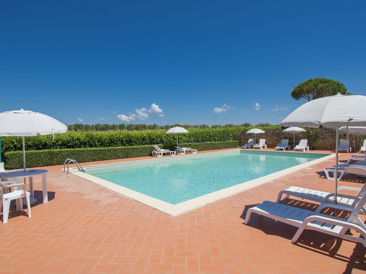 A rectangular swimming pool with clear blue water, surrounded by a tiled patio with lounge chairs and umbrellas.