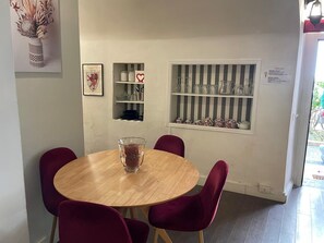 A round wooden table with four chairs, a glass vase with dried flowers, a framed picture, a shelf with kitchenware, and a window with striped blinds. - The Burgundy Nest by Campo de Fiori (Roma)