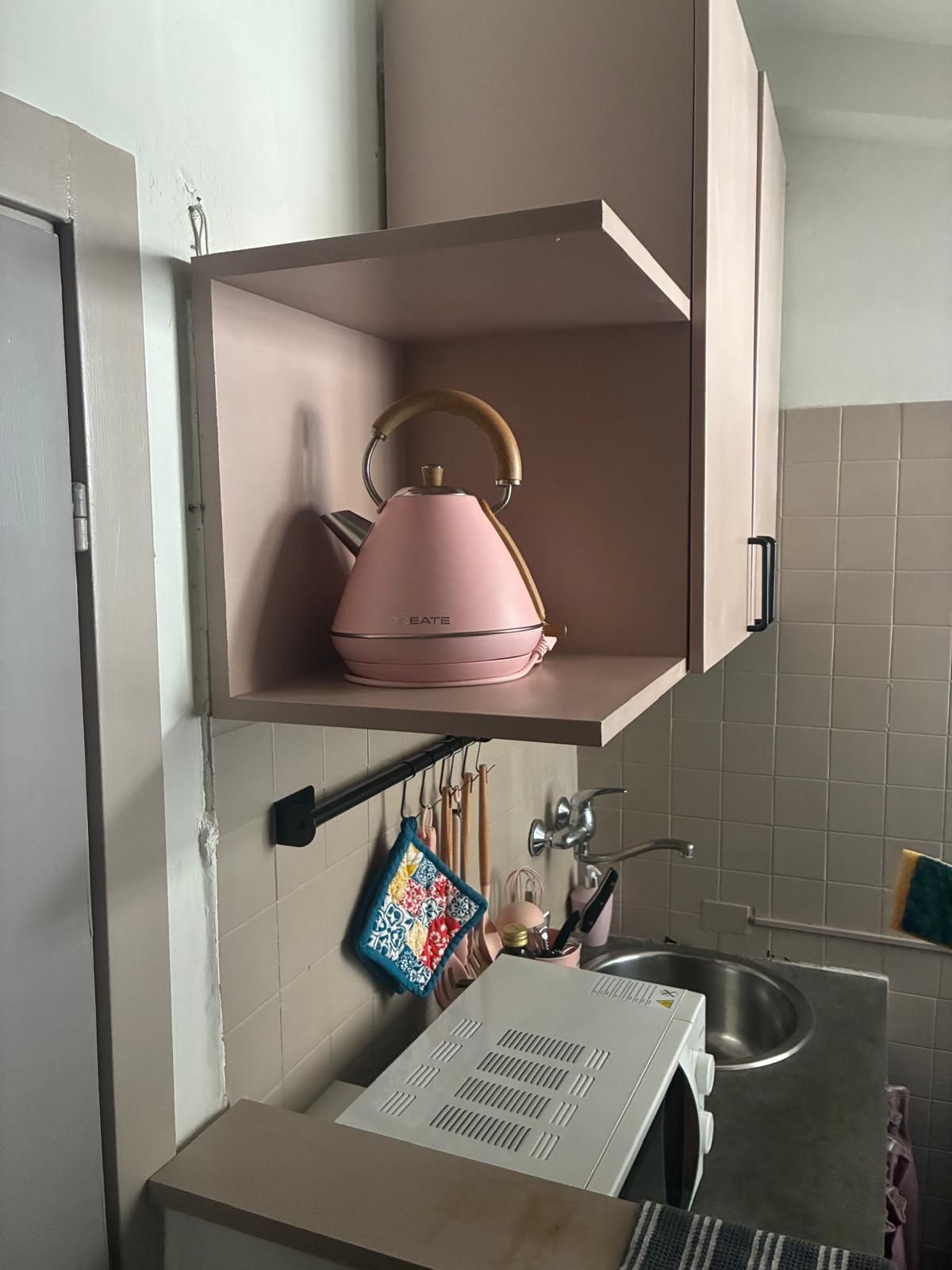 A kitchen with a pink kettle on a shelf, a hanging rack with kitchen utensils, and a sink.