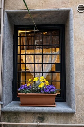 A window with a black metal grid, a flower box with yellow and purple flowers, and a curtain partially visible through the window. - The Burgundy Nest near Campo de Fiori (Roma)