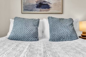 Een bed met een gebreide witte spreideken en twee vierkante kussens met een geweven patroon.