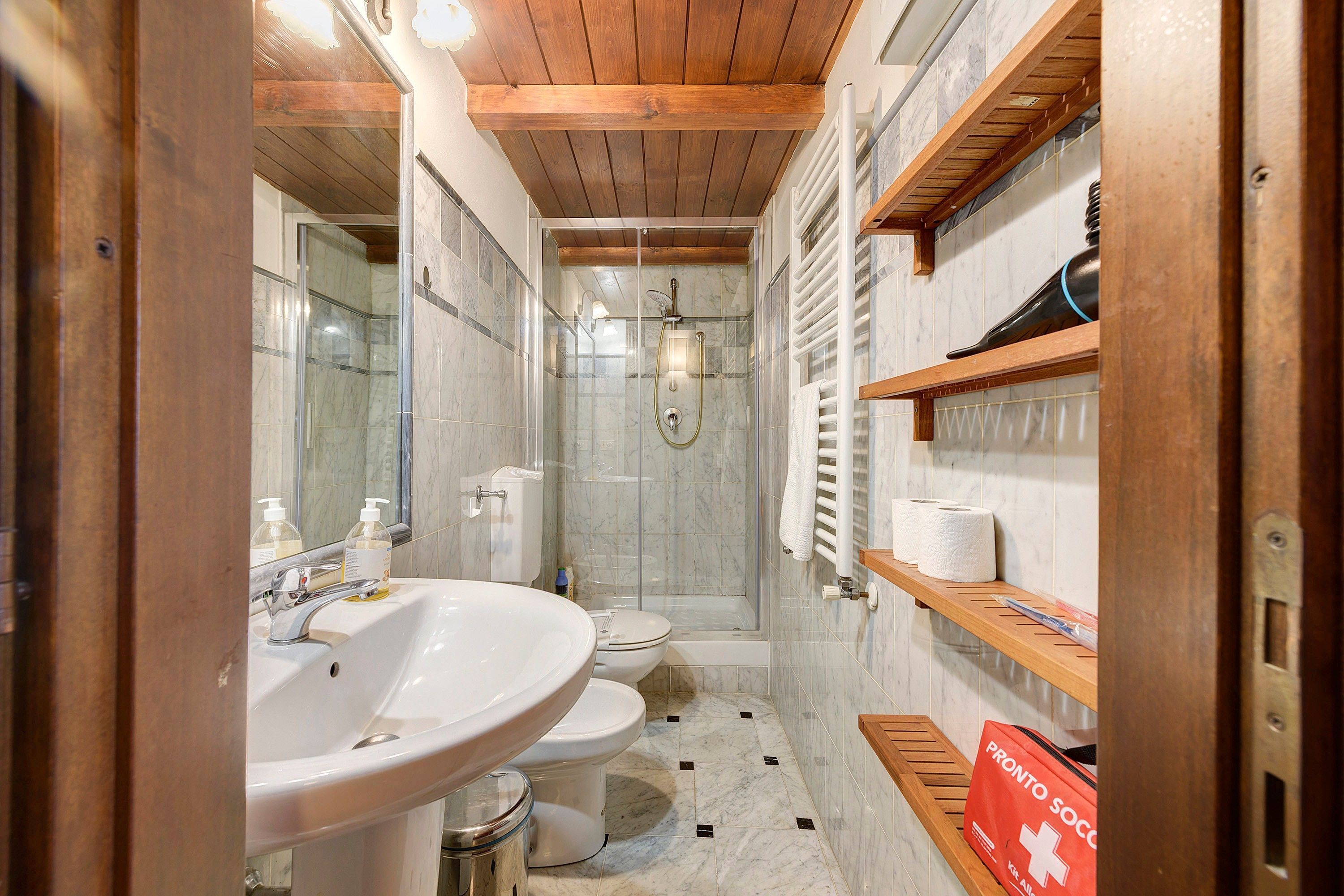 A bathroom with a wooden ceiling, a white sink, a toilet, a glass-enclosed shower, and a shelf with toiletries.
