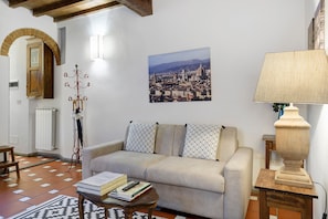 A living room with a sofa, a lamp, a coat rack, a radiator, and a cityscape painting on the wall. - Giulia Suite: Florentine Home in the Heart of Florence (Firenze)