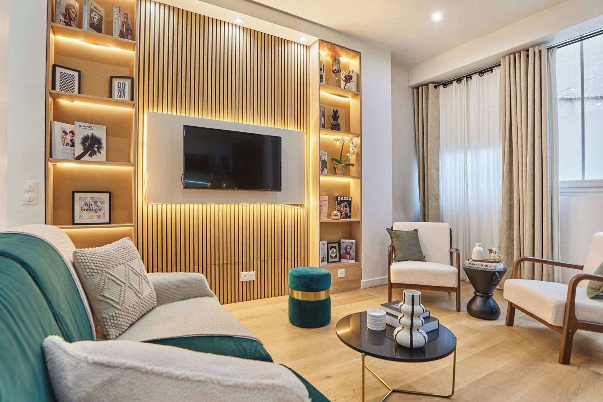 Un salon moderne comprenant un téléviseur à écran plat, un canapé, deux fauteuils, une table basse et une bibliothèque agrémentée d’objets décoratifs.
