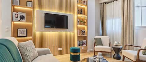 Un salon moderne comprenant un téléviseur à écran plat, un canapé, deux fauteuils, une table basse et une bibliothèque agrémentée d’objets décoratifs.