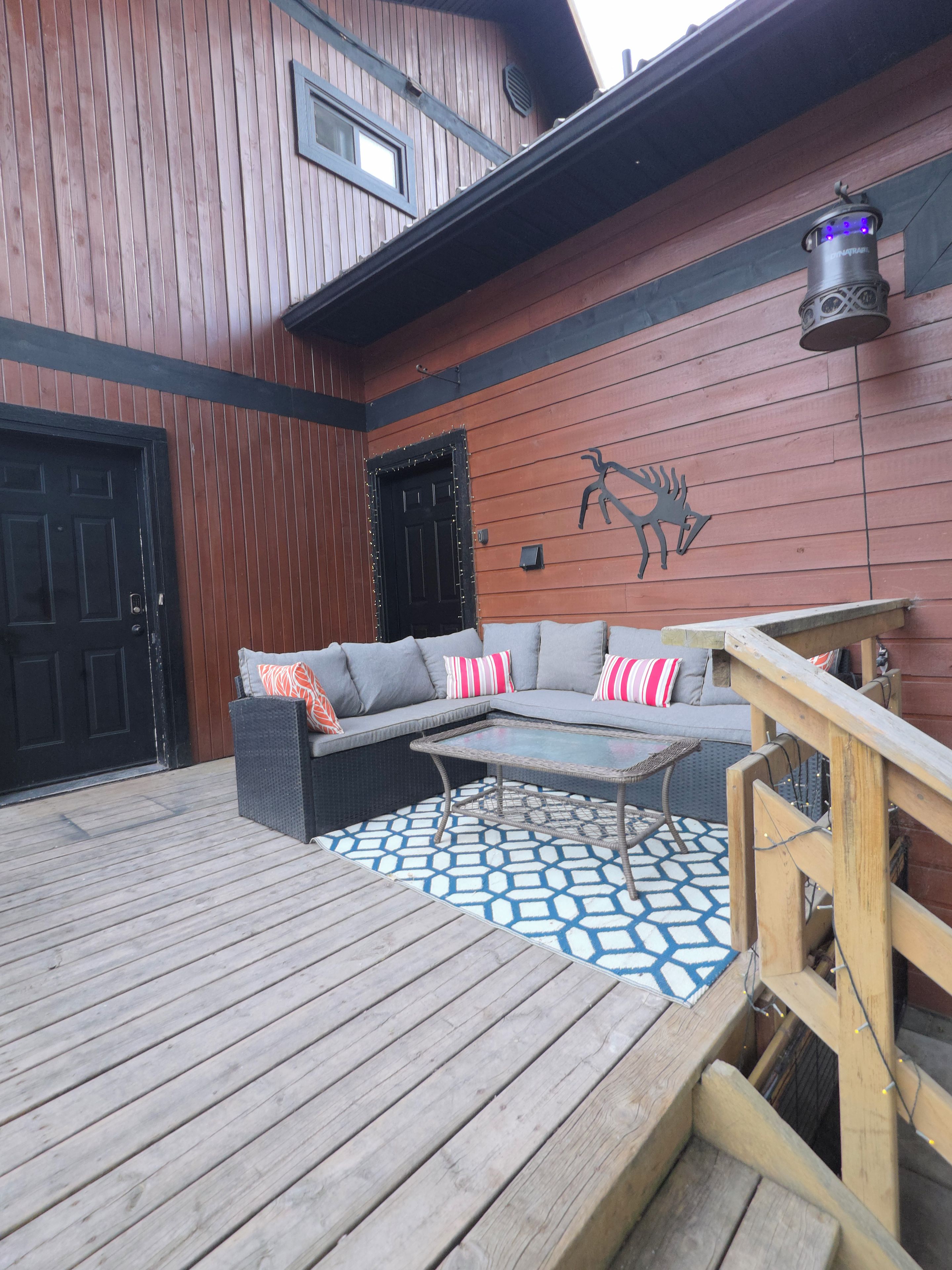 A wooden deck with a sofa and a table, a black door, and a wall-mounted light fixture.